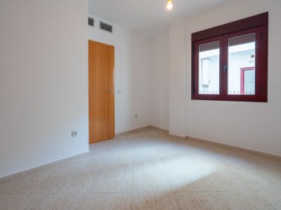 Empty room with light tiled floor and white walls. Features a wooden door and a dark red-framed window, looking out to an interior patio with a water heater. AC vents are visible above the door.