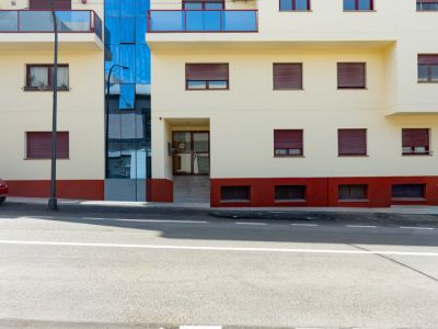 Exterior view of the residential building, featuring cream facade, blue glass balconies, and red roller shutters. A prominent central blue glass section and a street-level entrance with a ramp are visible.