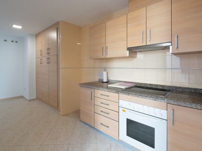 Functional kitchen featuring light wood-effect cabinetry, granite-effect countertop, integrated ceramic hob, and white oven. Tall pantry unit and beige floor tiles are visible.