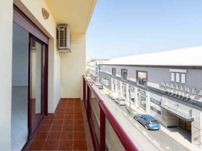 Outdoor balcony featuring terracotta floor tiles and a red railing. An air conditioning unit is visible, offering views of the street and "Martinez Muebles" store.