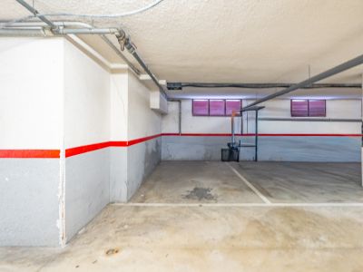 Private underground parking space with concrete floor, white and grey walls featuring a red stripe, and exposed ceiling pipes. Two small windows with red blinds are visible.