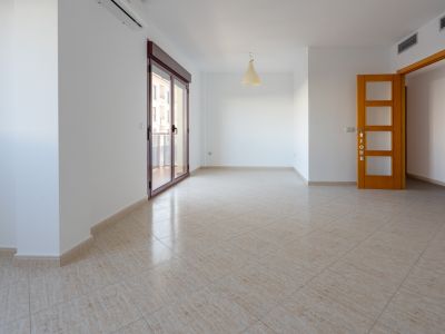 Spacious room with light beige ceramic tiled flooring. Features a large sliding glass door with a dark red frame to a terrace, a split AC unit, and a light wooden door with frosted glass panels.