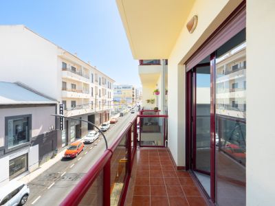 Sunny balcony featuring terracotta floor tiles and a red-framed glass railing, overlooking an urban street with buildings and traffic.