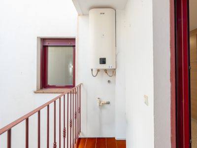 Utility patio featuring a wall-mounted Ferroli water heater, terracotta floor tiles, and a red metal railing. A burgundy-framed window with a granite sill and a matching door frame are visible.