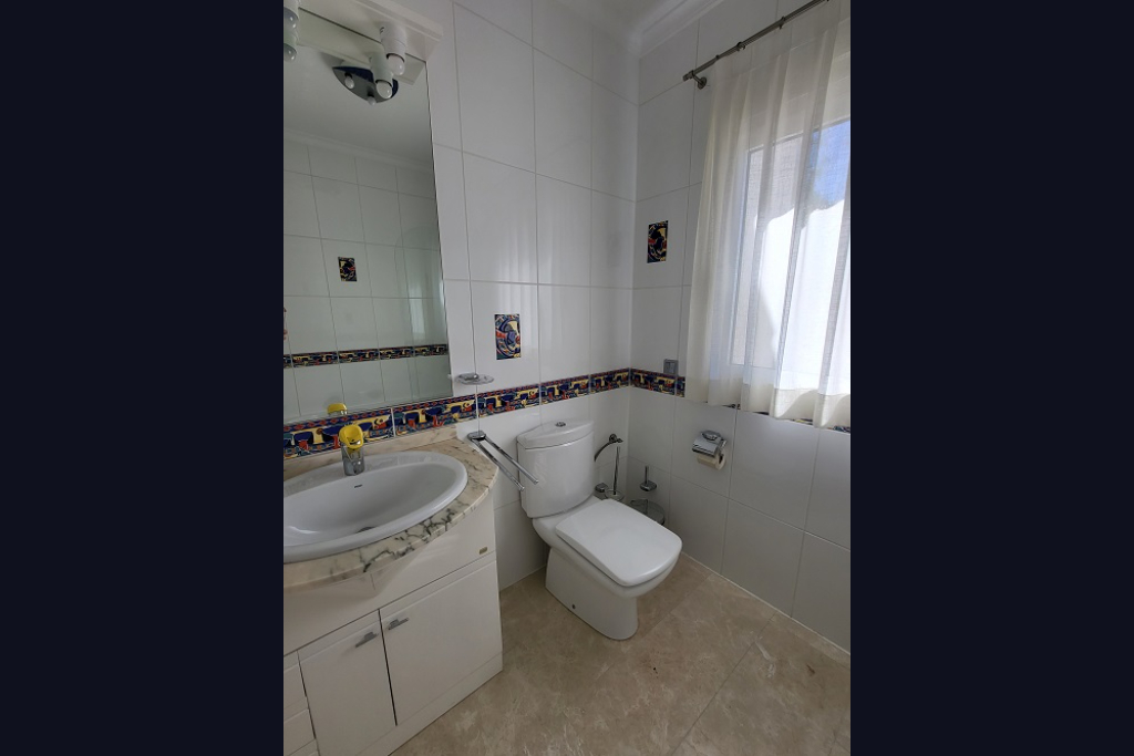 Bathroom featuring white tiled walls with a decorative border and rooster accents. Includes a sink with a light marble vanity, toilet, and window with sheer curtains.