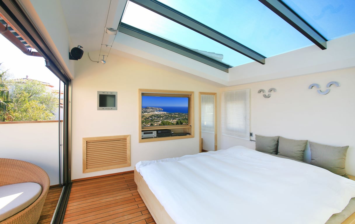 Bedroom featuring a large glass skylight and warm wooden flooring. A sliding glass door opens to a private terrace with a wicker chair, while a built-in TV displays coastal views.