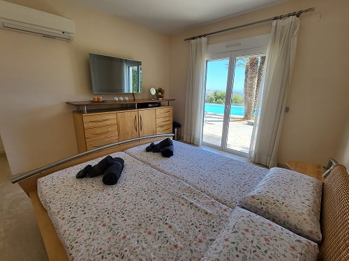 Bedroom featuring twin beds, a wall-mounted TV, and direct terrace access with pool and palm tree views. Includes air conditioning and wooden furniture.