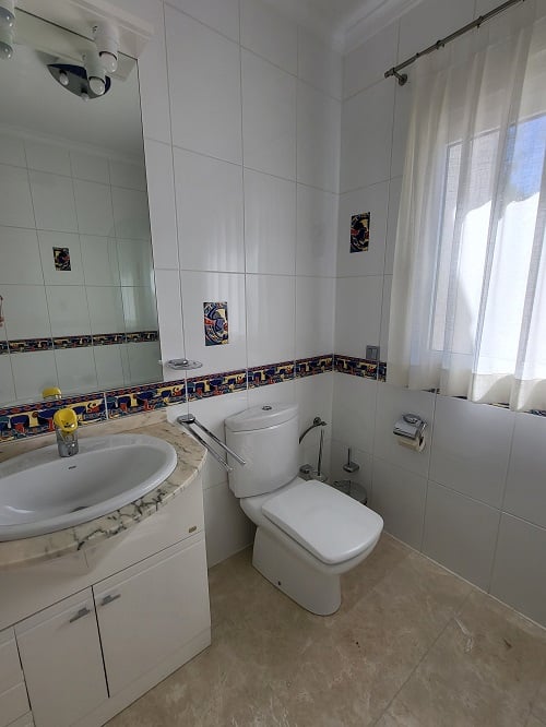 Bright bathroom featuring white wall tiles with a colorful decorative border and accent pieces. Includes a marble-topped vanity with integrated sink, a modern toilet, and a window with a sheer curtain.
