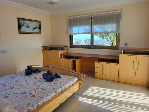 Bright bedroom featuring a light wood bed, integrated desk, and cabinetry. Windows offer panoramic coastal views, illuminating the space with natural light.