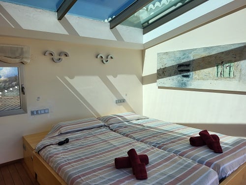 Bright bedroom featuring two single beds with striped linens and red towels. A prominent skylight bathes the room in natural light, complemented by an abstract painting and a window.