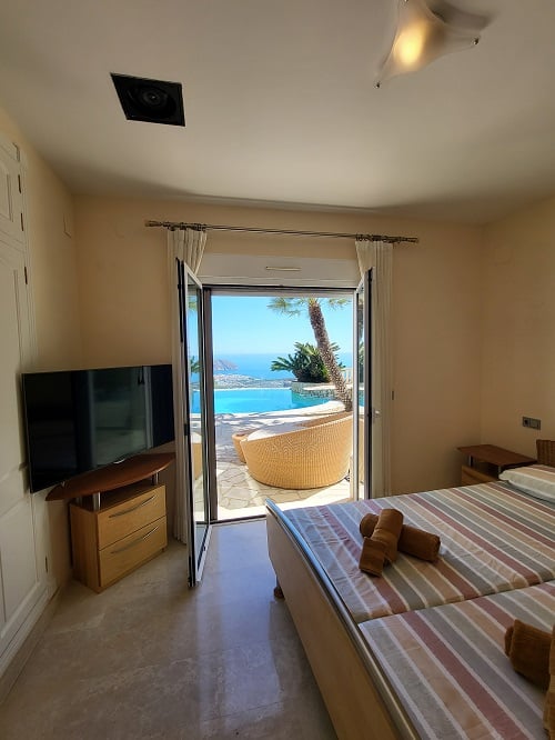 Bright bedroom with direct terrace access, featuring twin beds, a television, and light marble flooring. Offers panoramic views of the infinity pool and the Mediterranean Sea.