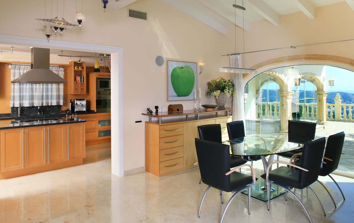 Bright dining area with a glass table and black chairs, featuring polished marble floors. Large arched windows frame panoramic sea and mountain views, adjacent to a modern kitchen.
