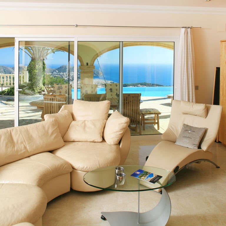 Bright living area featuring light curved sofas and marble flooring, with direct access to a terrace showcasing an infinity pool and panoramic sea views.