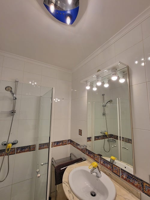 Compact bathroom featuring white tiled walls with a decorative border, a glass-screened shower, and a marble-effect vanity with an illuminated mirror. A unique blue and silver ceiling light adds a modern touch.