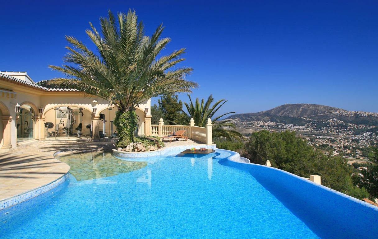 Curved mosaic pool with palm island and submerged table. Glass-enclosed gym and terrace offering panoramic mountain views.