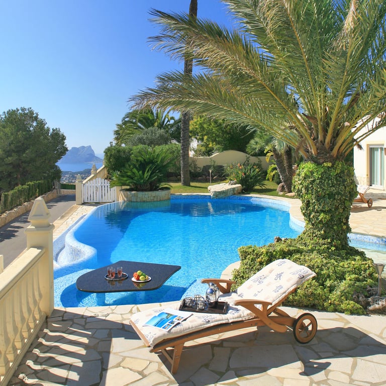 Curved swimming pool featuring a submerged black table with refreshments, adjacent to a stone-paved terrace with a sun lounger. Distant sea views and a prominent rock formation are visible under a clear sky.