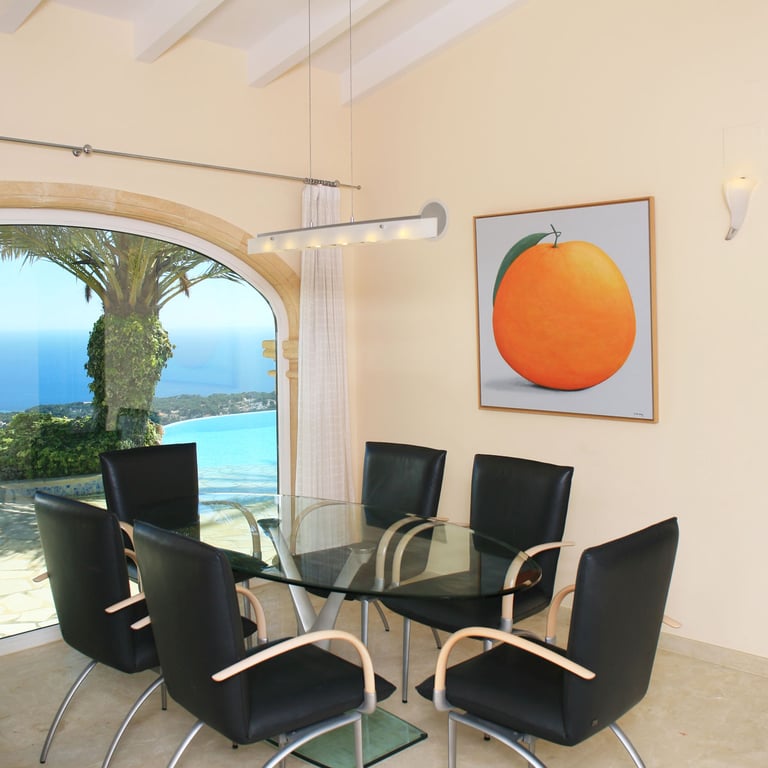 Dining area featuring a glass table and modern chairs, complemented by a large arched window with panoramic sea and coastline views. A prominent orange artwork adorns the wall.