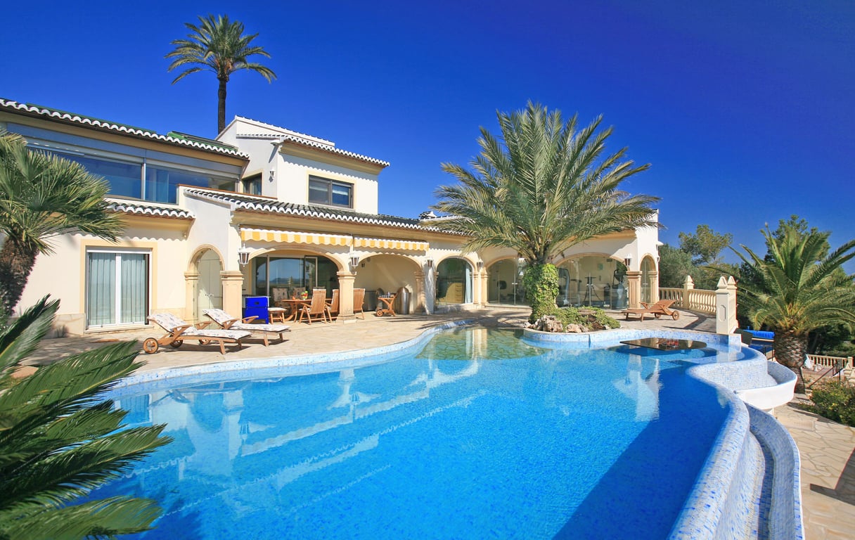 Expansive blue mosaic pool with beach-style entry, palm island, and in-water table. The villa features a covered naya and a glass-fronted gym.