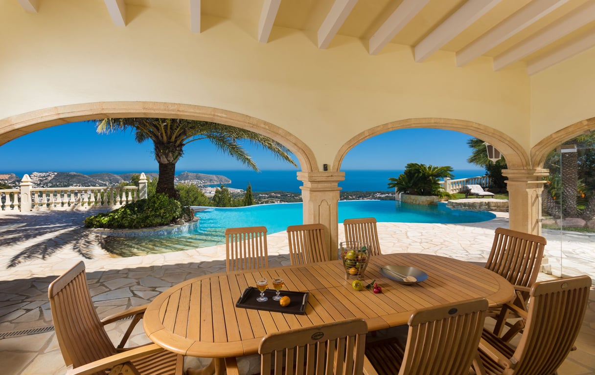 Expansive covered terrace featuring a wooden dining table and chairs, framed by stone arches revealing panoramic Mediterranean sea and coastal views with an infinity pool.