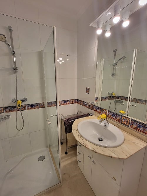 Functional bathroom featuring a glass shower cabin, vanity with light marble countertop, and an illuminated mirror. White tiles with a decorative Mediterranean border.