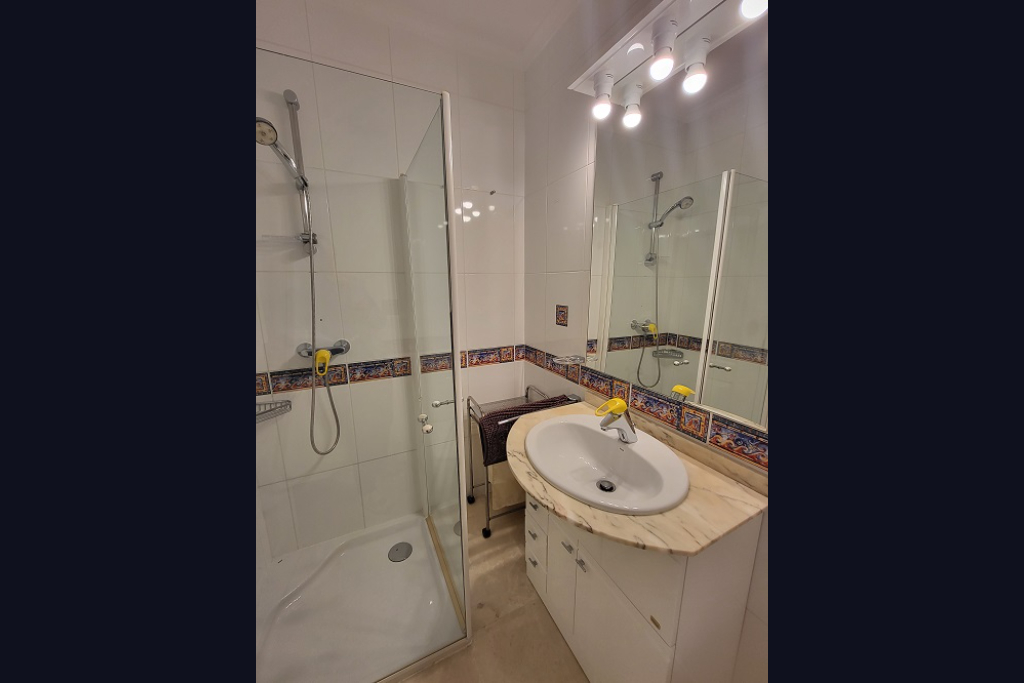 Functional bathroom featuring a walk-in shower with a glass screen, a basin set in a light marble-patterned countertop, and a white vanity. White wall tiles are accented with a decorative Mediterranean-style border.