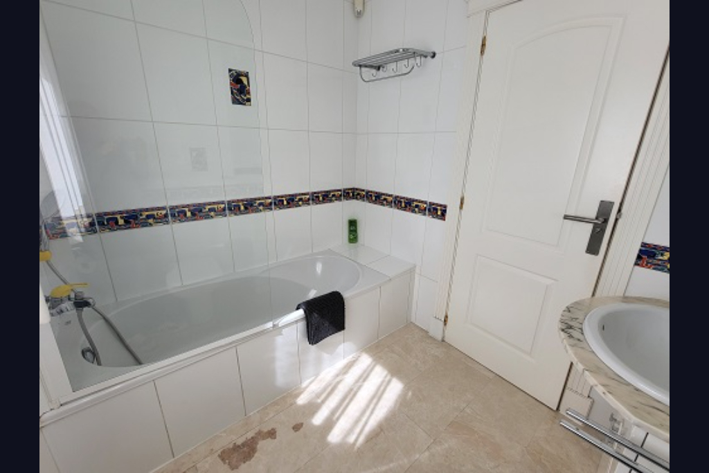 Functional bathroom with a white bathtub, glass screen, and walls tiled in white with a decorative border. Features a marble-topped sink and natural light.
