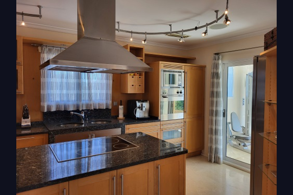 Modern kitchen featuring light wood cabinetry, dark granite countertops, an island with integrated hob, and stainless steel appliances. A glass door leads to an adjacent gym space.