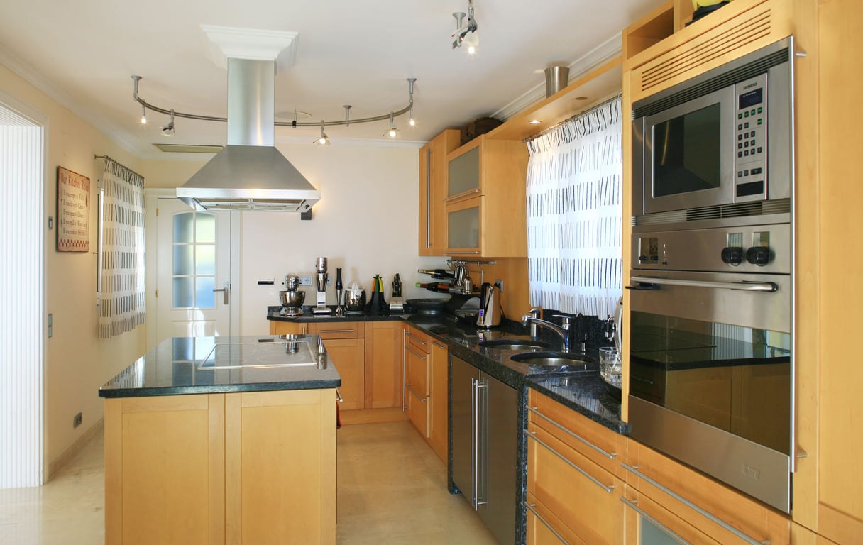 Modern kitchen featuring light wood cabinetry, dark granite countertops, and a central island with an integrated hob. Stainless steel appliances include a built-in oven, microwave, and extractor hood.