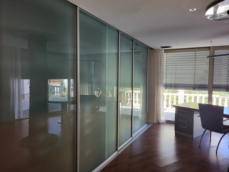 Office or bedroom featuring sleek frosted glass sliding doors, dark wood flooring, and a dedicated workspace by the window overlooking the balustrade.