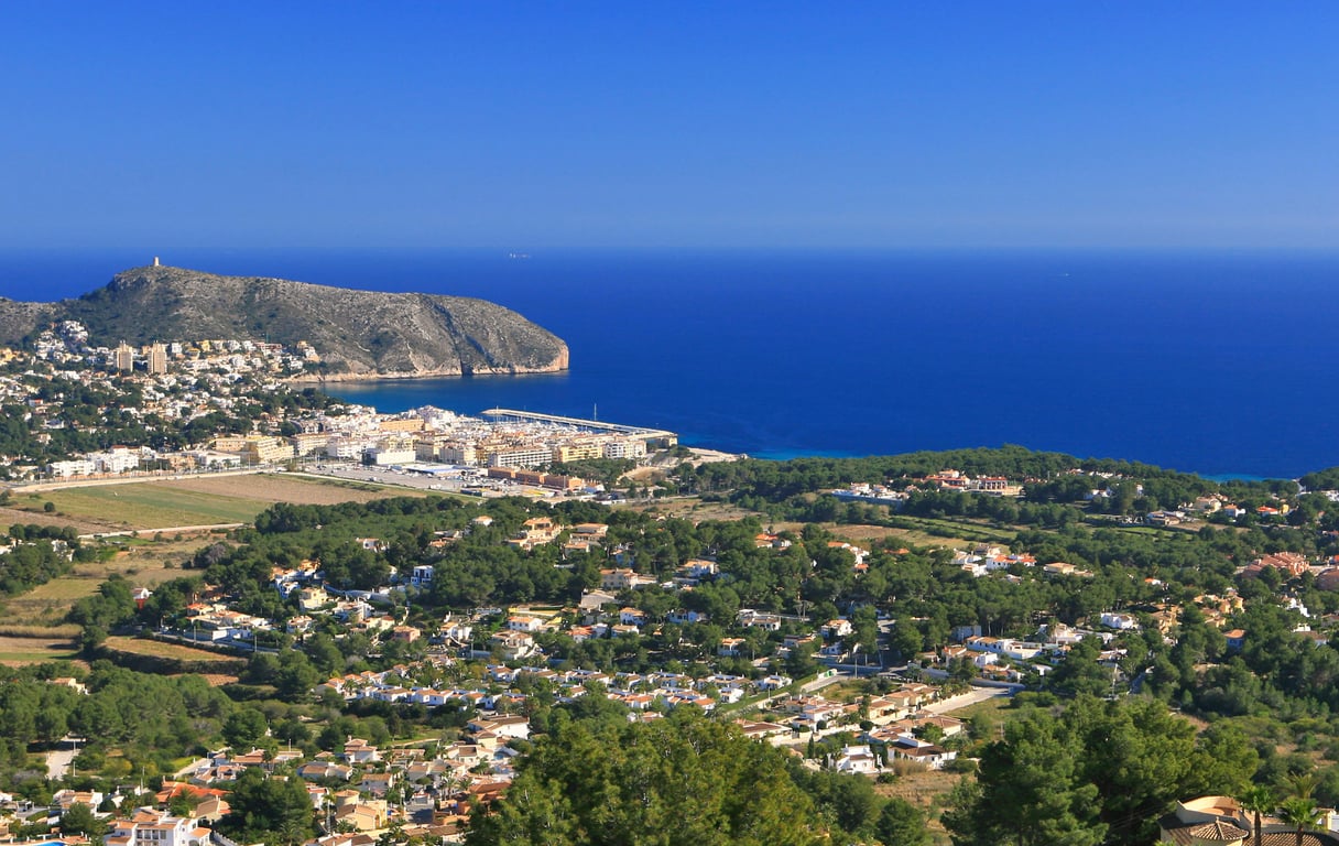 Panoramic view from a hillside, featuring a coastal town, a marina, a prominent mountain, and the deep blue Mediterranean Sea under a clear sky.