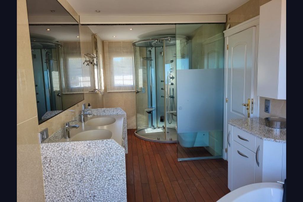 Spacious bathroom featuring a double vanity with pebble-textured base, a circular hydro-massage shower, and warm wooden flooring. Large mirror and windows with blinds are visible.