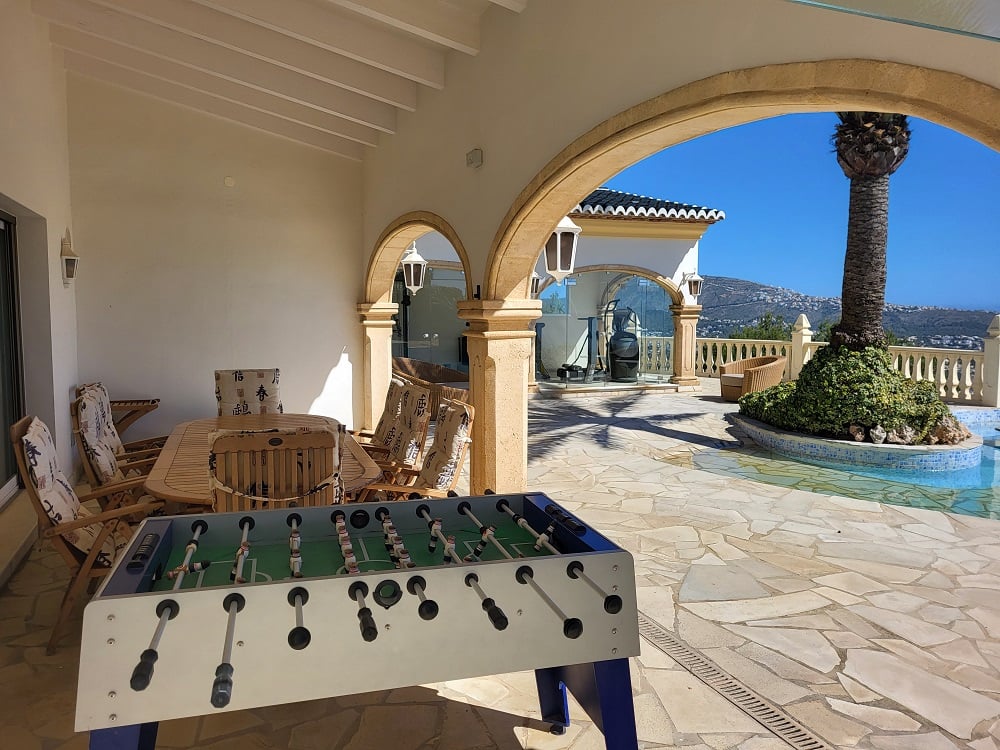Spacious covered naya featuring a wooden dining set and a foosball table, framed by stone arches and irregular flagstone flooring. Overlooks the pool with a palm island and distant mountains.