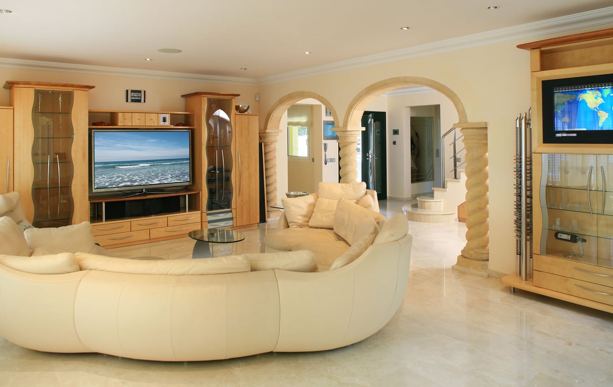Spacious living room featuring a large curved cream sofa, light marble floors, and custom light wood entertainment units. Arched stone columns lead to the entrance and staircase.
