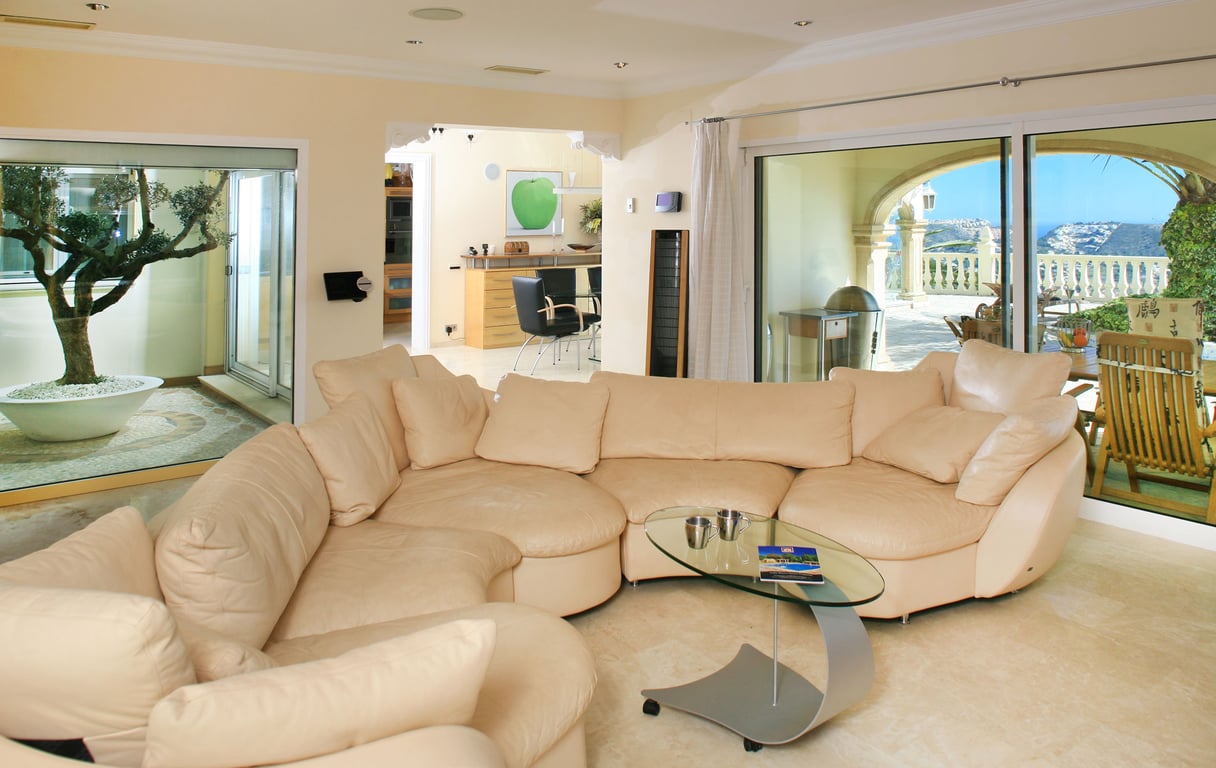 Spacious living room featuring a light sectional sofa, glass coffee table, and marble floors. Large windows offer views to a sea-facing terrace and an internal olive tree courtyard.