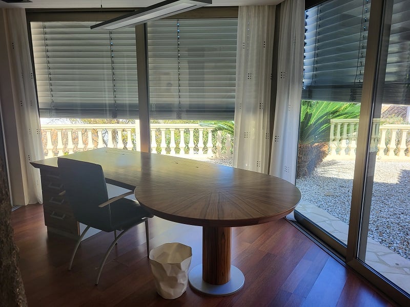 Spacious private office featuring a large wooden desk and hardwood floors. Floor-to-ceiling windows overlook a balustrade and Mediterranean garden.