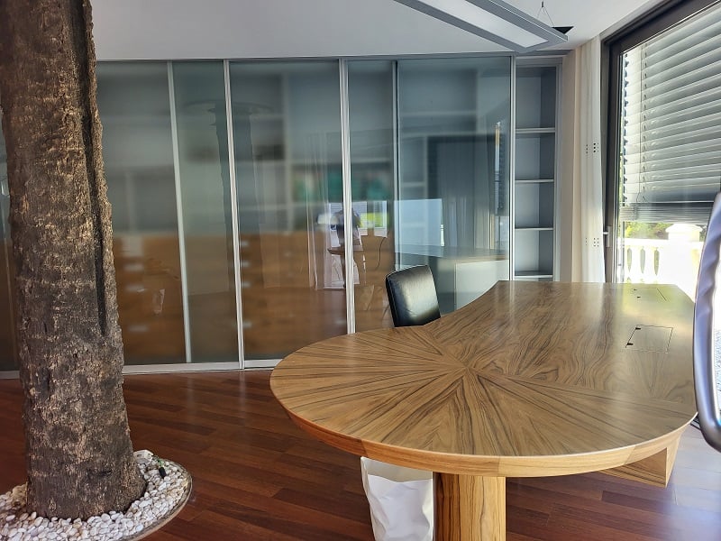 Spacious private office featuring a unique wooden desk with a starburst pattern, frosted glass sliding doors, an indoor tree, and natural light from a large window.