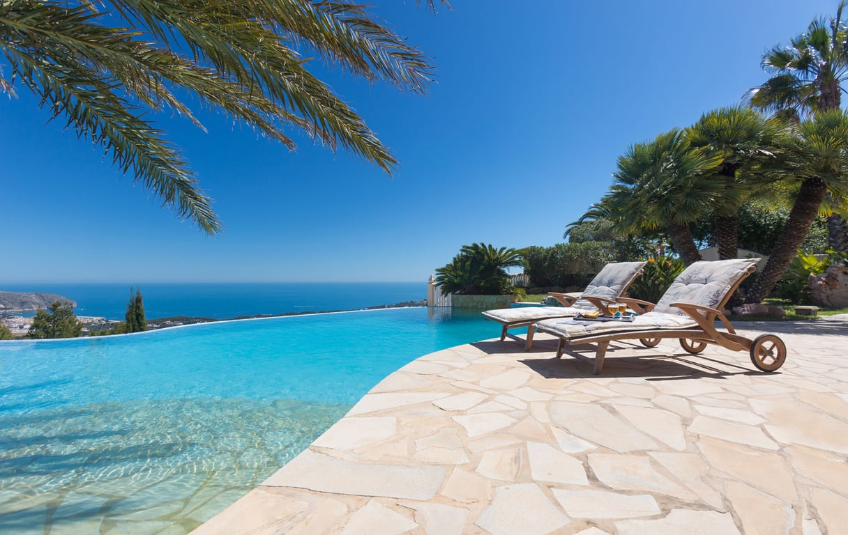 The infinity pool terrace features two wooden sun loungers on a flagstone patio, offering expansive views of the azure Mediterranean Sea and distant coastline under a clear sky.