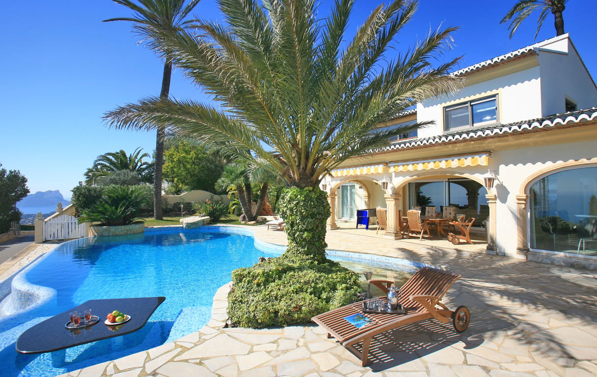 The uniquely designed pool features an in-water table for refreshments, surrounded by a stone terrace and palm trees, with distant sea views.