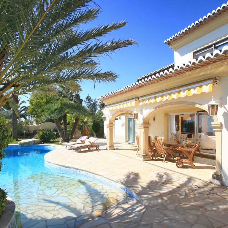 The villa's exterior features a freeform swimming pool and a spacious stone-paved terrace leading to a covered naya with a wooden dining set, all under striped awnings.