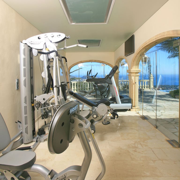 This private gym features Hoist and Life Fitness equipment, marble flooring, and arched glass walls offering panoramic sea and pool views. Skylights provide natural light.
