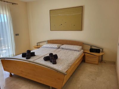 Bedroom featuring a wooden bed frame with a woven headboard and metal accents. Bedside tables with drawers flank the bed, beneath an abstract painting. Natural light enters through sheer curtains.
