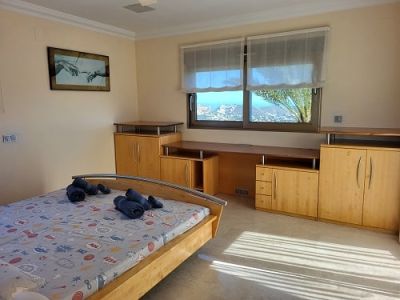 Bright bedroom featuring a light wood bed, integrated desk, and cabinetry. Windows offer panoramic coastal views, illuminating the space with natural light.