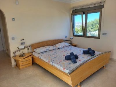 Bright bedroom featuring a light wooden bed, woven headboard, and a nightstand. The open window frames views of lush greenery and distant mountains.