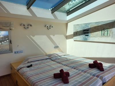 Bright bedroom featuring two single beds with striped linens and red towels. A prominent skylight bathes the room in natural light, complemented by an abstract painting and a window.