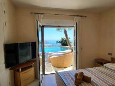 Bright bedroom with direct terrace access, featuring twin beds, a television, and light marble flooring. Offers panoramic views of the infinity pool and the Mediterranean Sea.