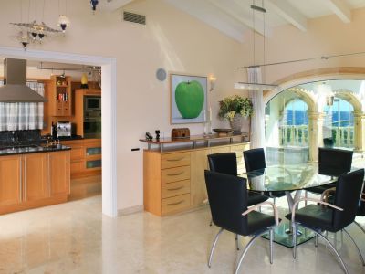 Bright dining area with a glass table and black chairs, featuring polished marble floors. Large arched windows frame panoramic sea and mountain views, adjacent to a modern kitchen.