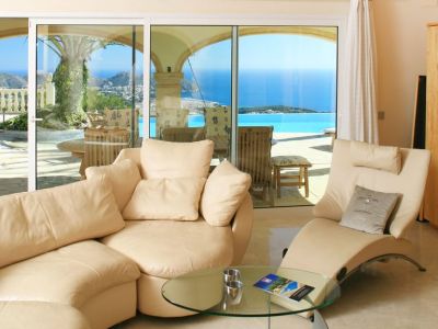 Bright living area featuring light curved sofas and marble flooring, with direct access to a terrace showcasing an infinity pool and panoramic sea views.