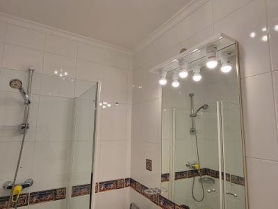 Compact bathroom featuring white tiled walls with a decorative border, a glass-screened shower, and a marble-effect vanity with an illuminated mirror. A unique blue and silver ceiling light adds a modern touch.