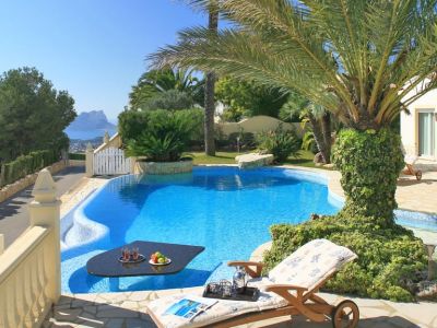 Curved swimming pool featuring a submerged black table with refreshments, adjacent to a stone-paved terrace with a sun lounger. Distant sea views and a prominent rock formation are visible under a clear sky.