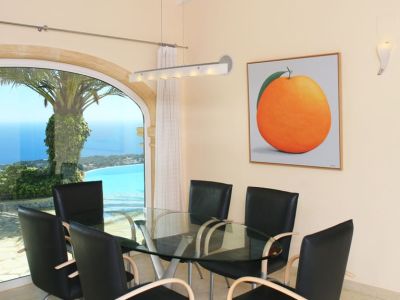 Dining area featuring a glass table and modern chairs, complemented by a large arched window with panoramic sea and coastline views. A prominent orange artwork adorns the wall.